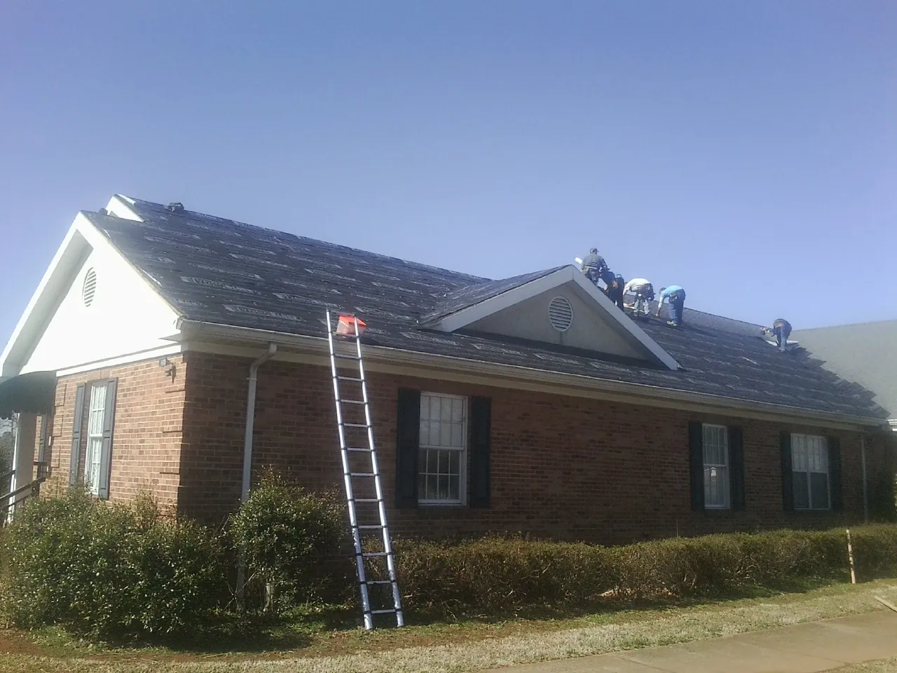 Crew tearing shingles off a brick ranch roof with a ladder set — Metal Roof Repair work by our crew in Genoa